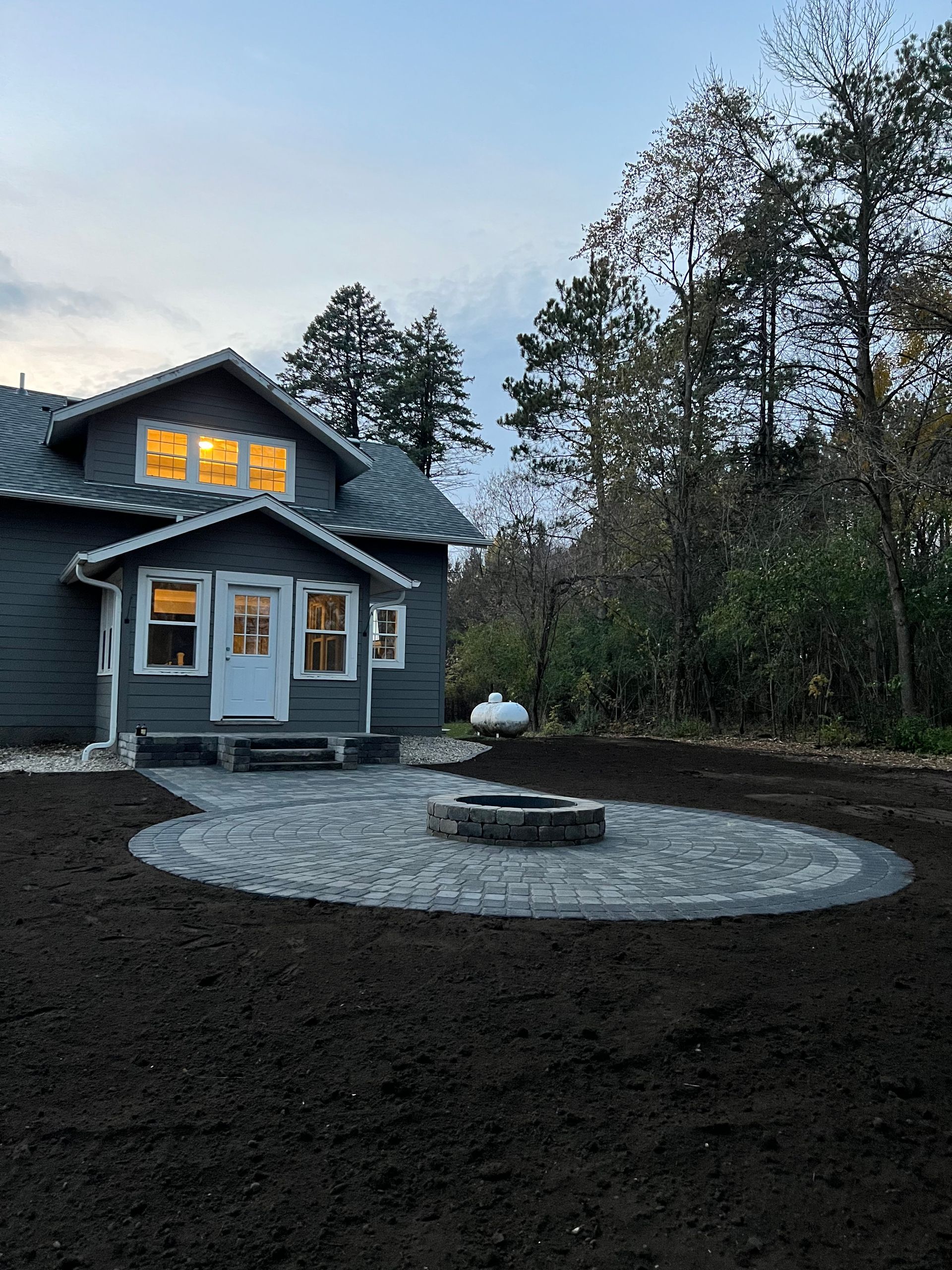 House with lit windows and a stone fire pit in a dark yard at dusk, surrounded by trees