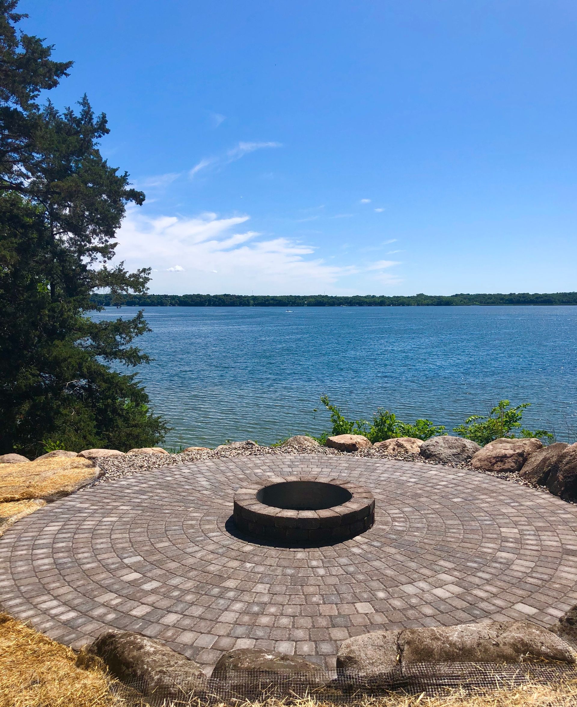 Stone fire pit on a cobblestone patio beside a calm blue lake under a clear sky.