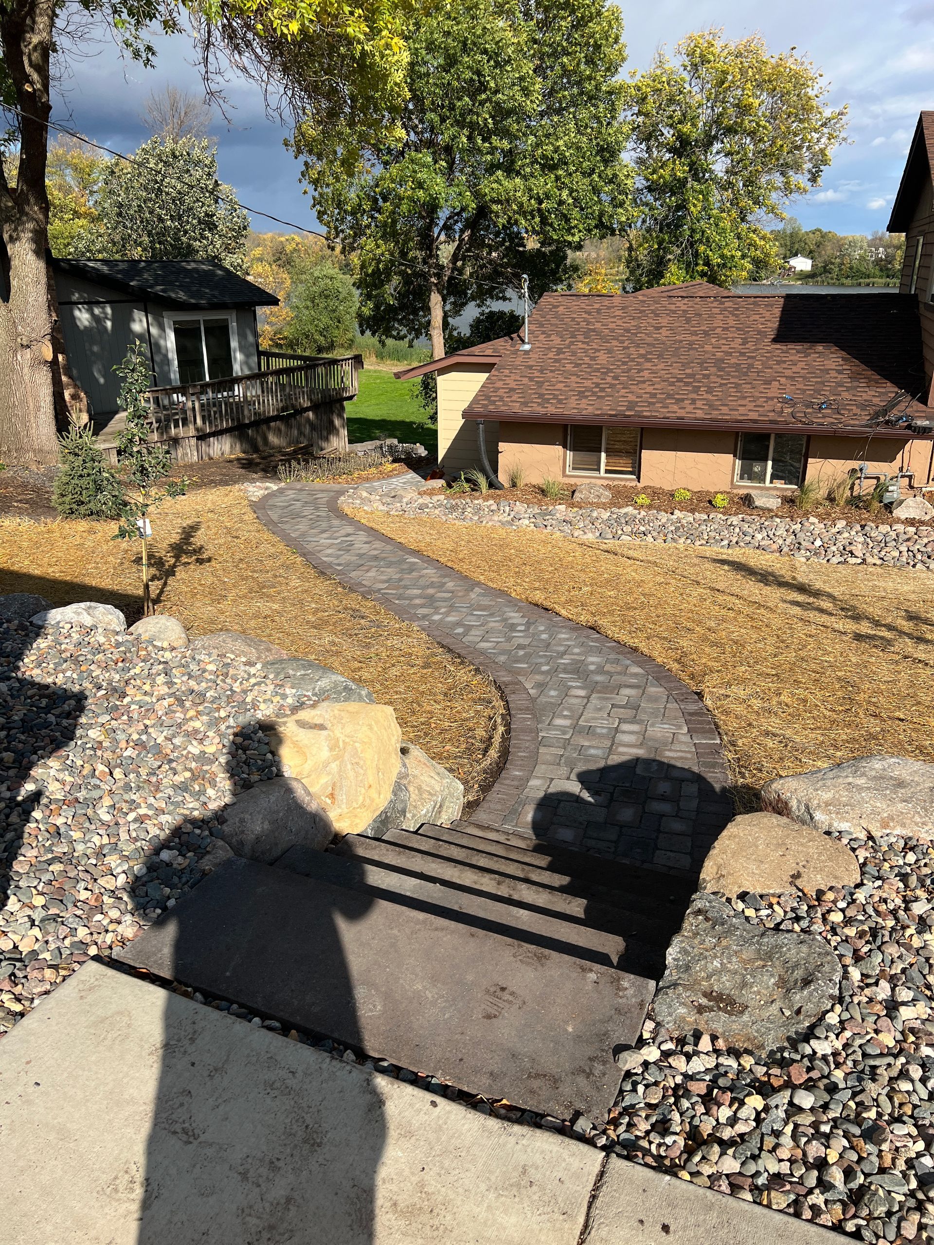 Garden path curving through gravel with rocks, trees, and small rustic sheds in sunlight