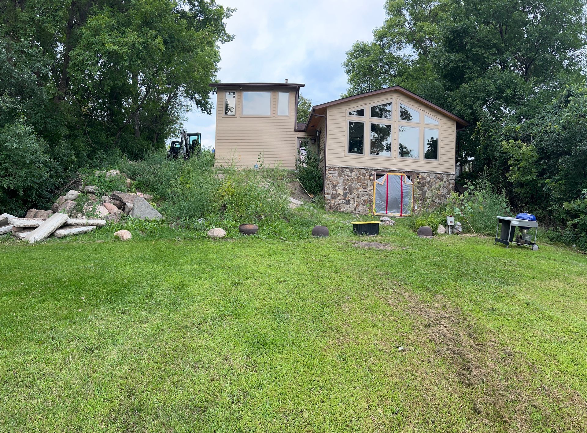 Rural yard with a small house, tall grass, scattered debris, and trees under an overcast sky