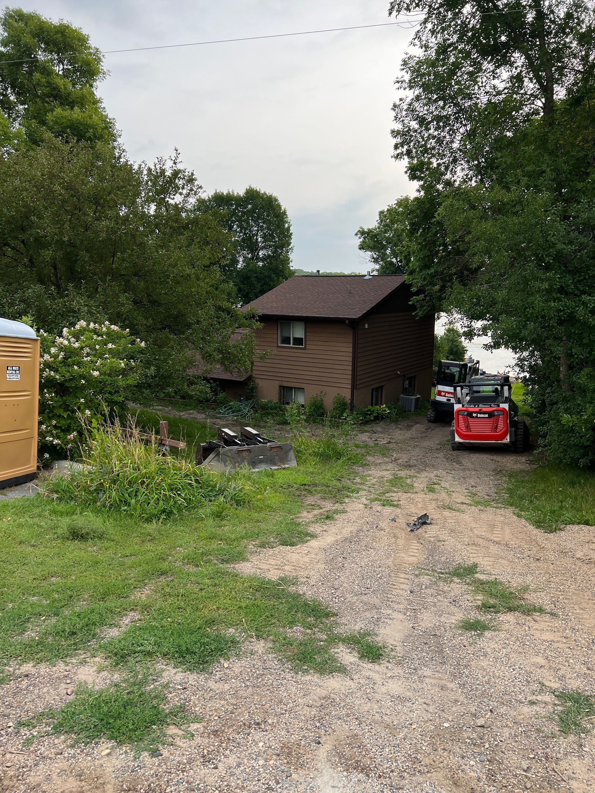 Gravel driveway with a brown shed, trees, and a red toy car near a portable toilet.