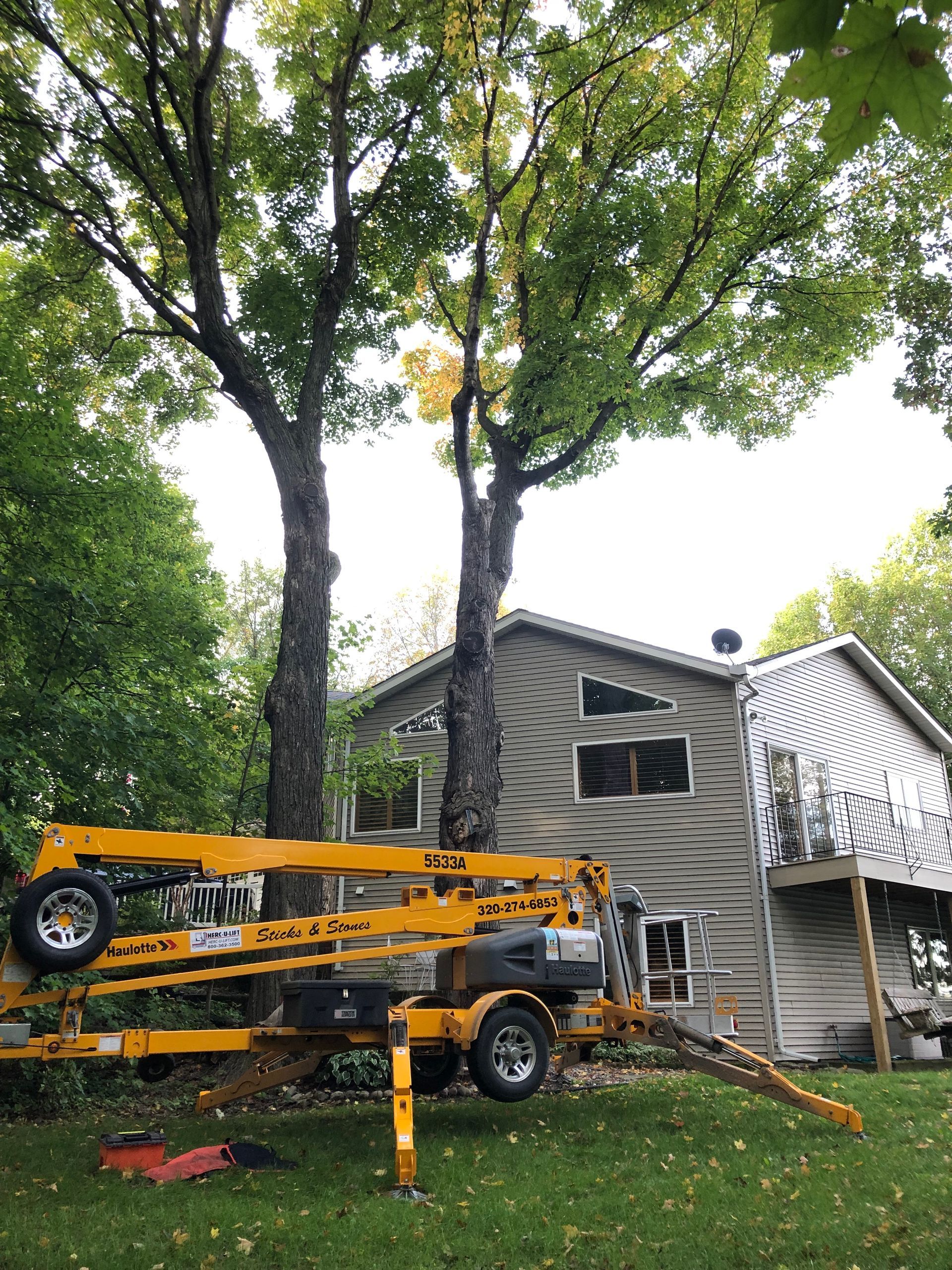Yellow boom lift parked in front of a gray house under large trees