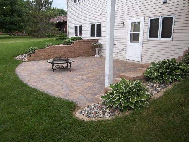 Back patio with paver walkway, fire pit, and hosta plants beside a white house