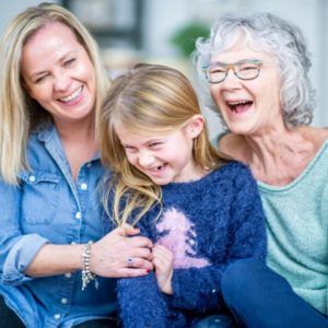 Three smiling people sit closely together indoors, laughing and embracing.