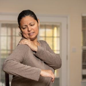 Woman holding her shoulder and elbow, wincing in pain indoors.