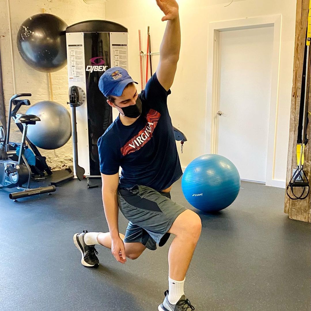Person lunging in a gym, wearing a mask and blue cap, with exercise balls in the background.