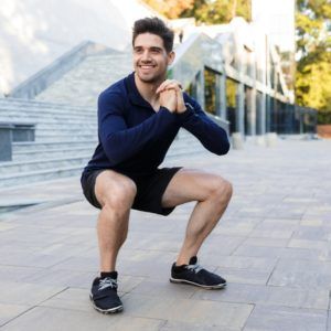Man doing a squat exercise outdoors on a paved plaza, wearing a navy shirt, black shorts, and sneakers