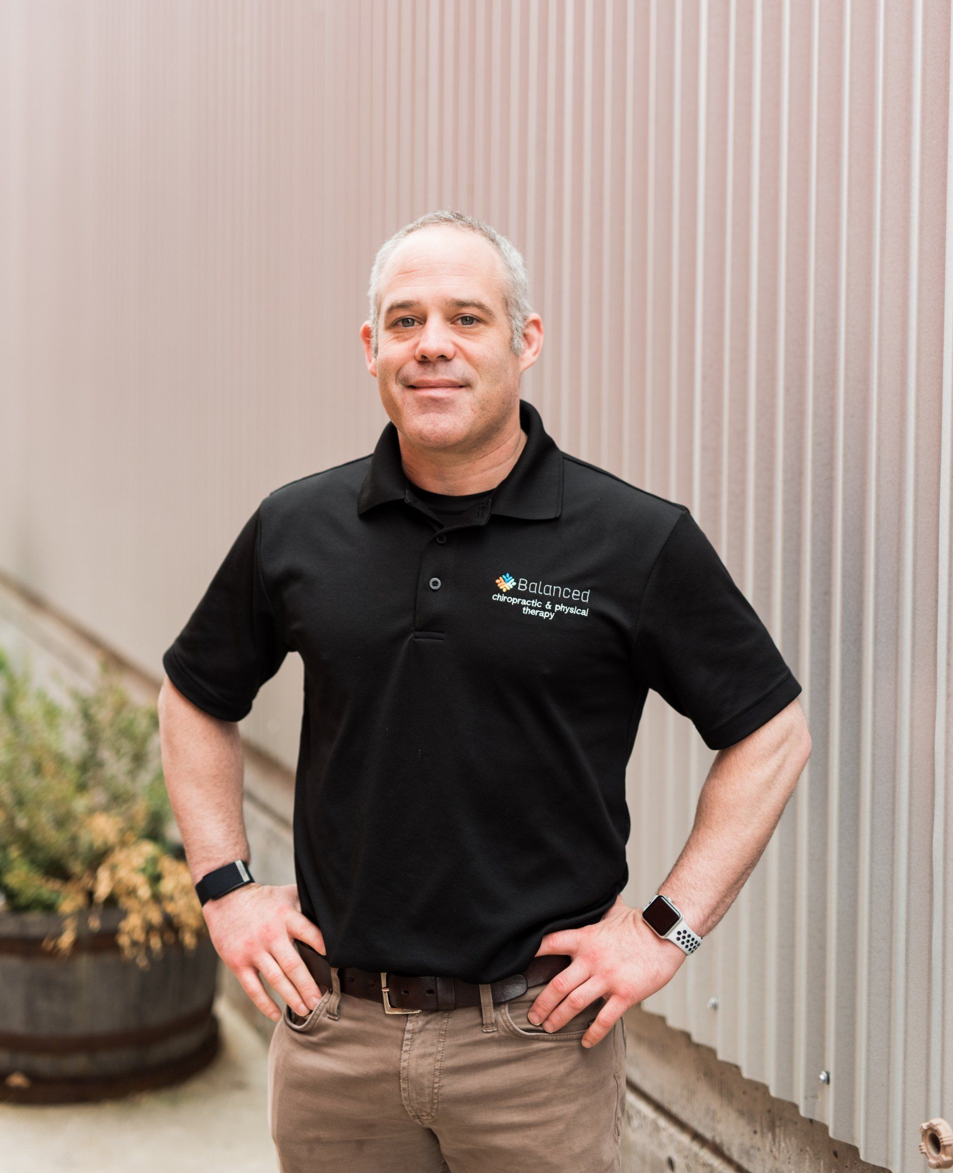 Man standing outdoors against a corrugated wall, wearing a black polo shirt and khaki pants.