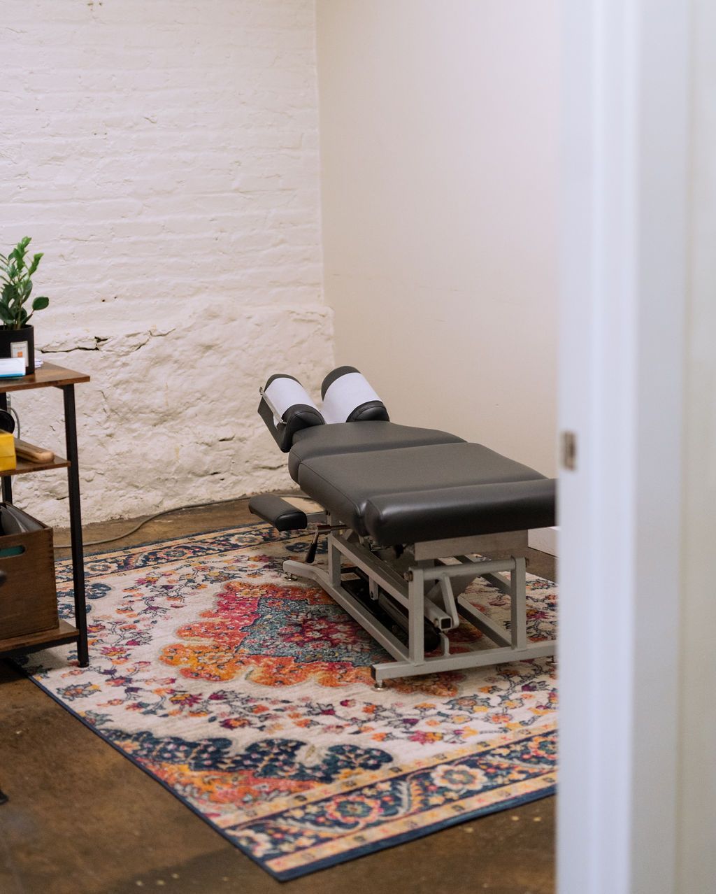 Adjustable black exam table with feet visible in a small treatment room on a patterned rug