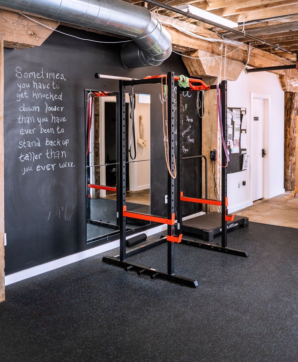 Home gym with squat rack, chalkboard wall, and exposed beams on rubber flooring.