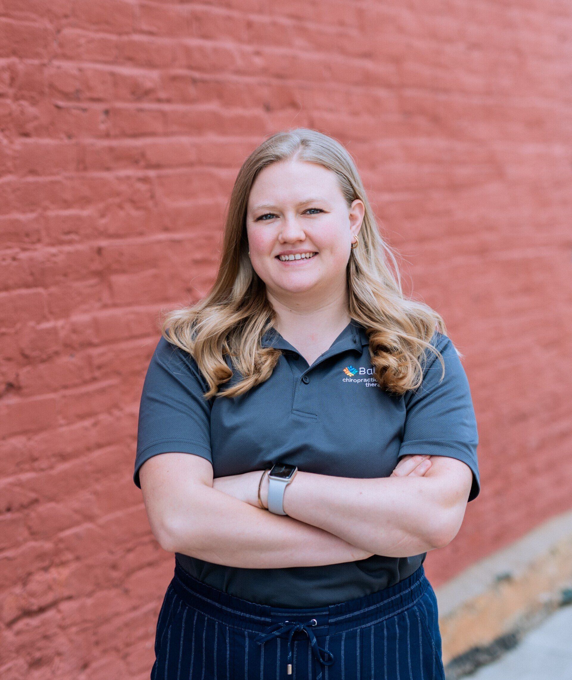 Smiling person with crossed arms in a navy shirt, standing against a red brick wall.