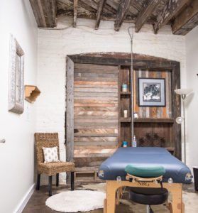 Rustic room with wood beams, sliding barn door, wicker chair, floor lamp, and blue massage table