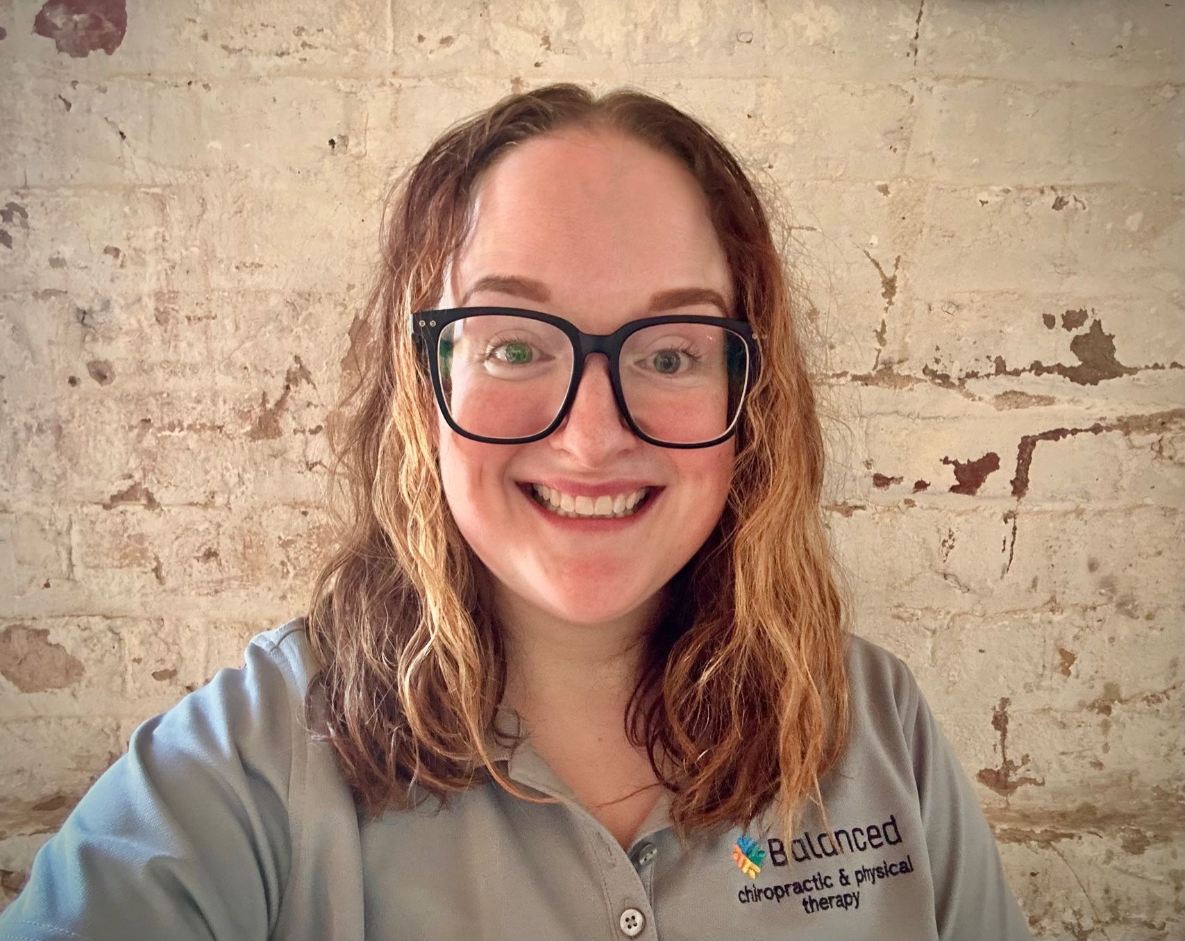 Smiling person with glasses and wavy hair in a gray logo shirt against a brick wall