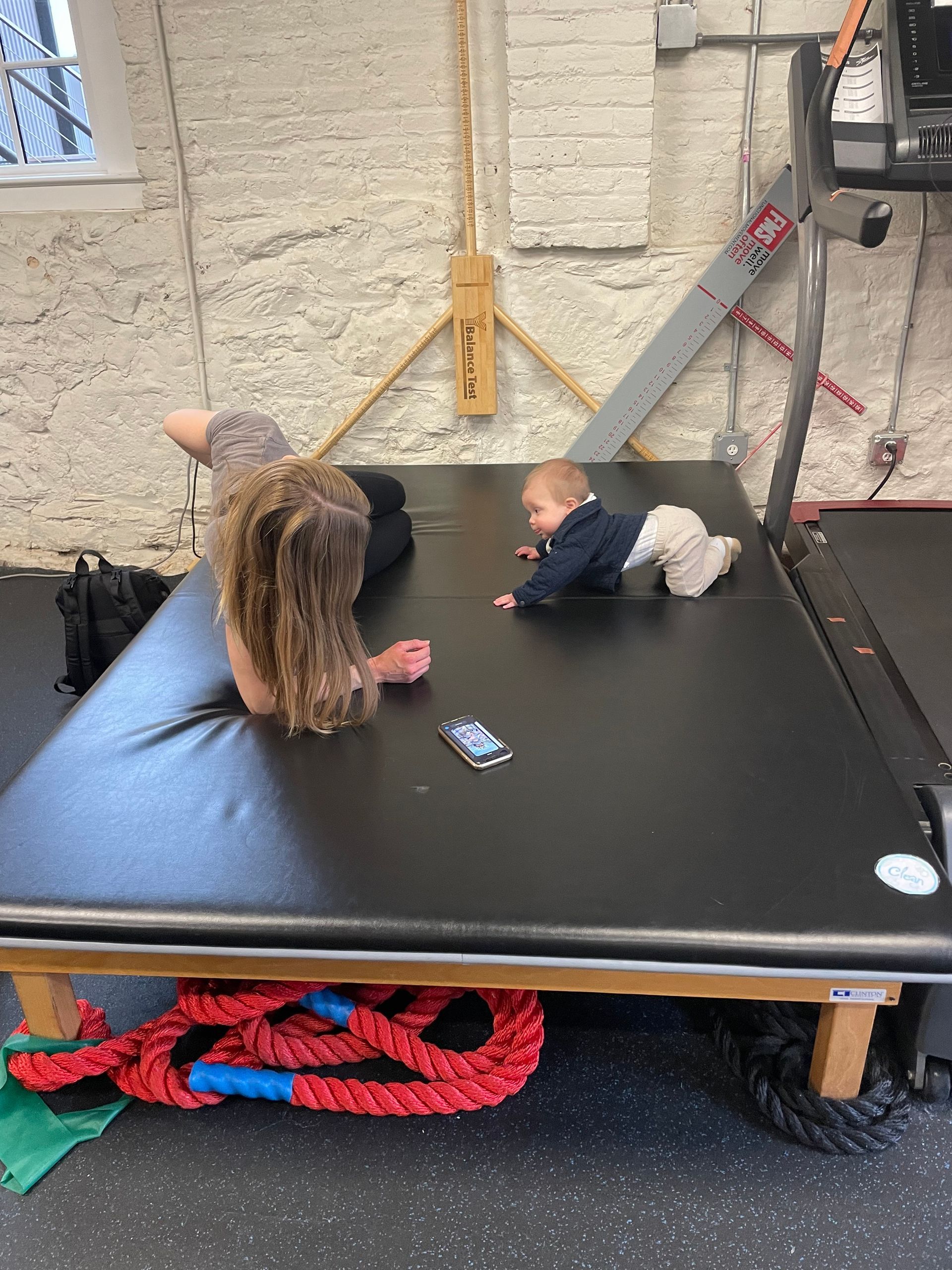 Two children lying on a black padded platform in a gym-like room, with ropes and equipment nearby