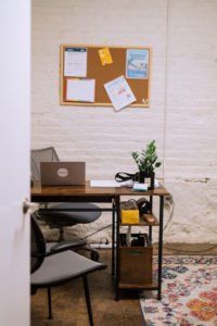 Small home office with desk, laptop, chair, wall corkboard, plant, and patterned rug on a white brick wall.