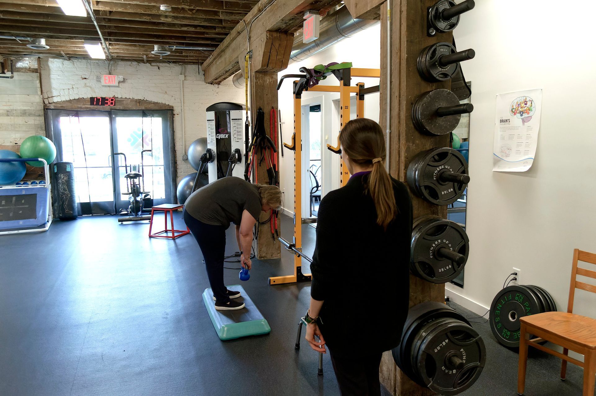 Person balancing on a step platform in a gym while another watches near weight racks