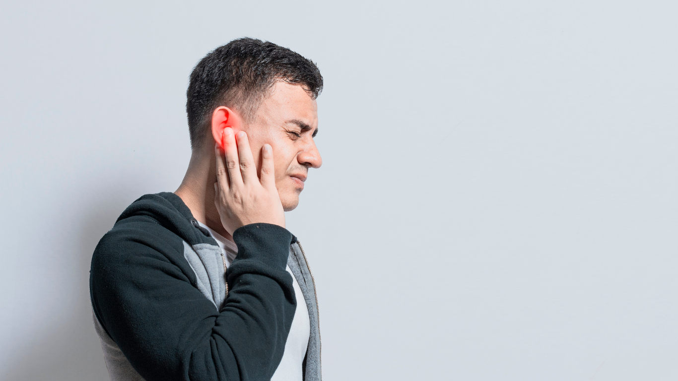 Man in gray sweater touching his ear and wincing against a light gray background
