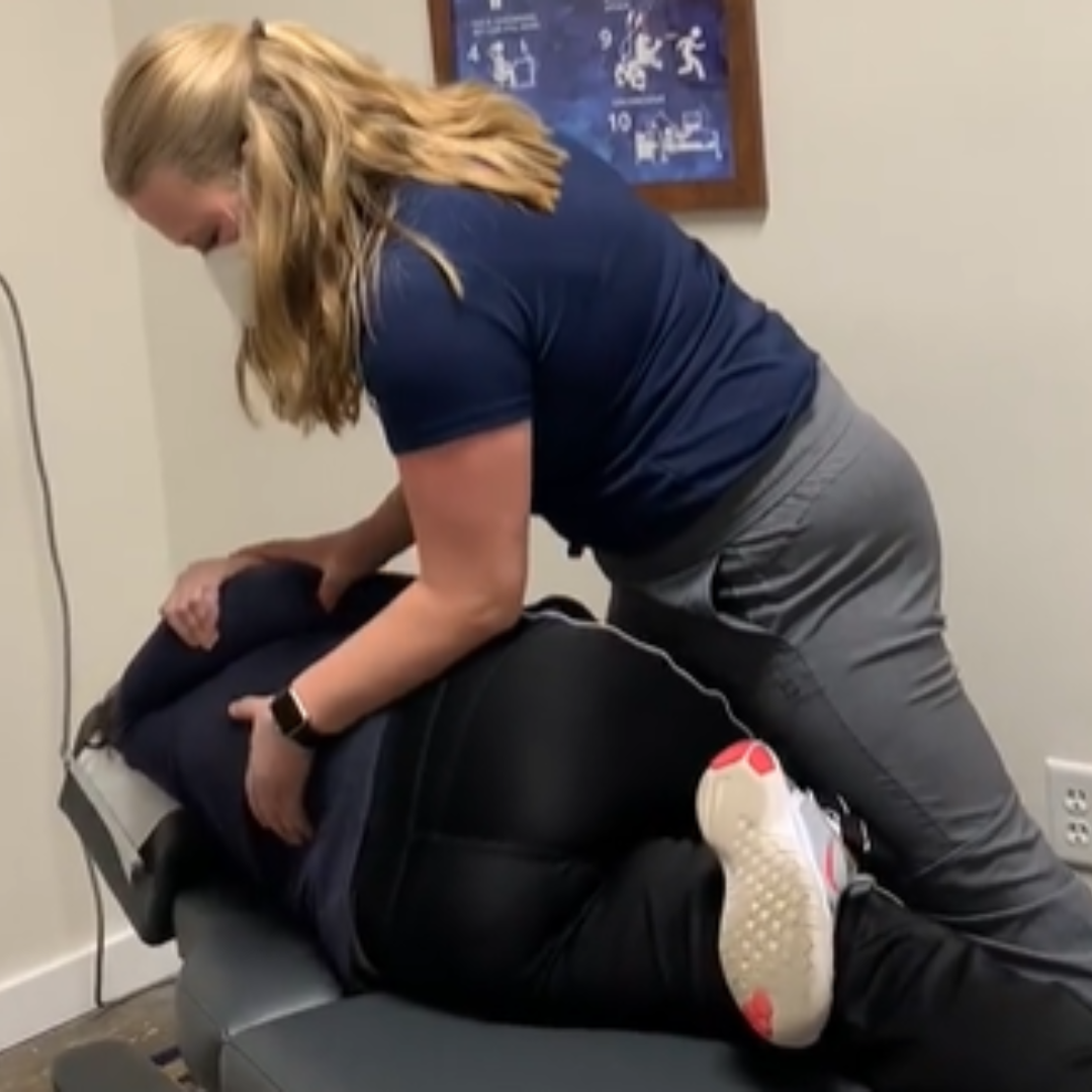 Woman in navy shirt and gray pants assisting a person on a treatment table in a clinic room