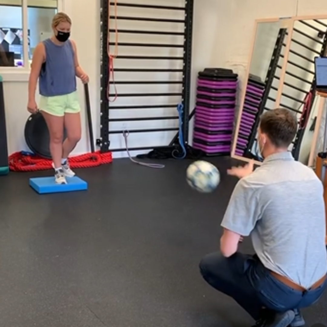 Two people exercising in a gym, balancing on foam pads and passing a ball.