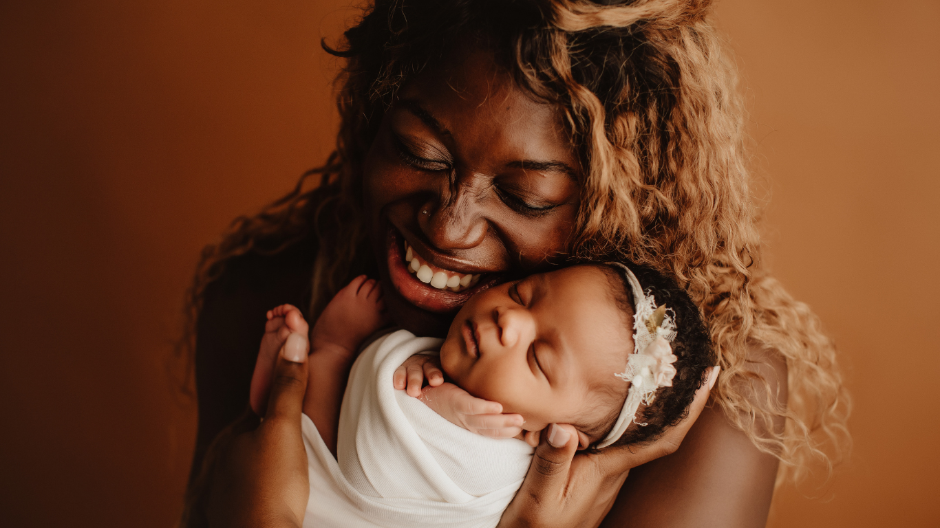 Smiling parent holding a sleeping baby wrapped in white against a warm brown background