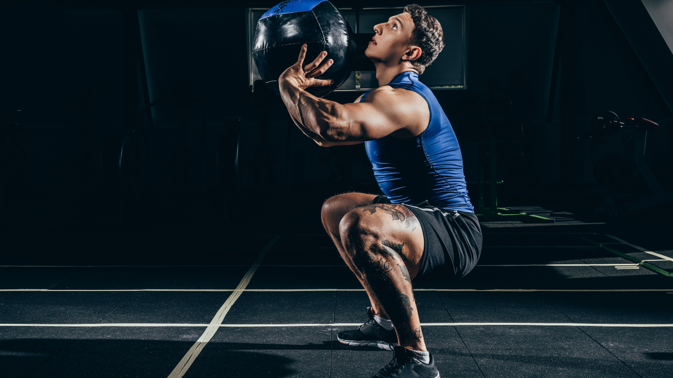 Athlete in blue shirt and black shorts holding a medicine ball in a dark gym, mid-squat exercise