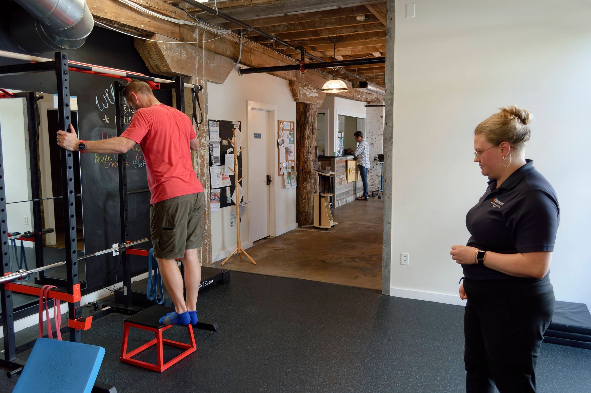 Two people in a gym; one stands on a box adjusting a wall-mounted bar while the other watches.