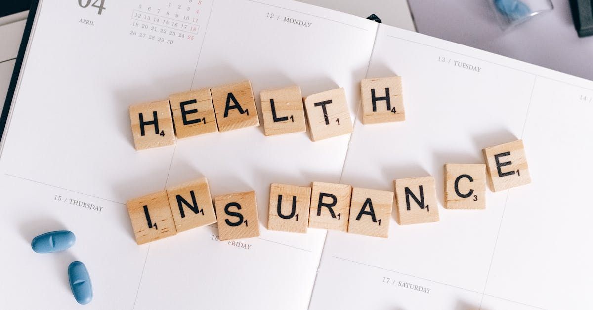 Scrabble tiles spelling “HEALTH INSURANCE” on a desk with papers and capsules nearby