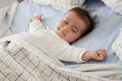 Sleeping baby in white pajamas lying on a bed with checkered blankets and a blue pillow