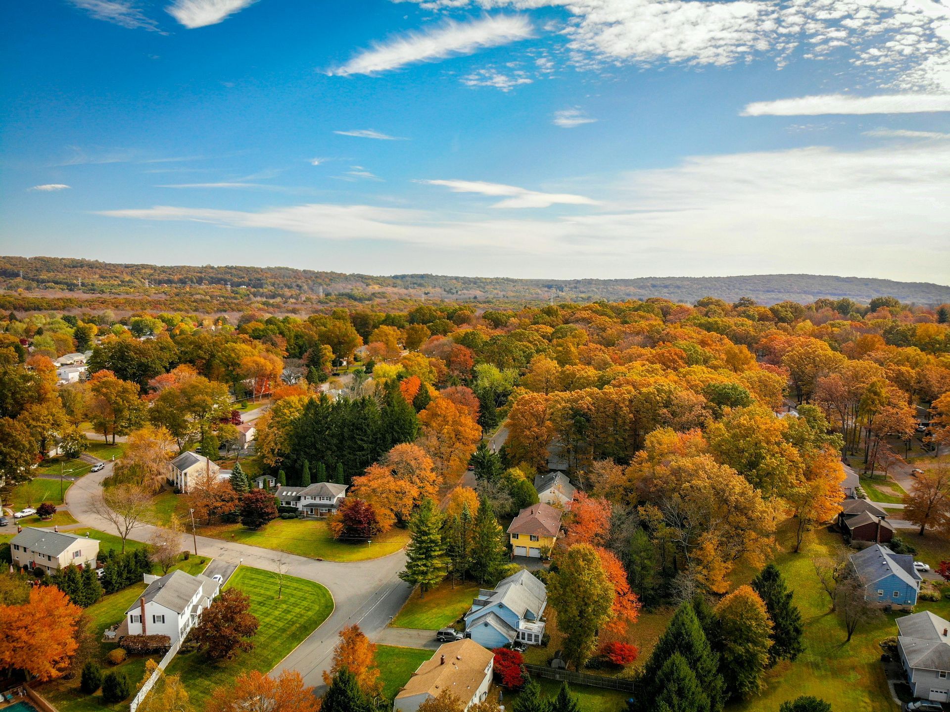 Aerial view of suburban homes surrounded by colorful autumn trees under a blue sky.
