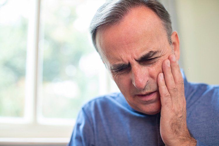 Man holding his cheek with a pained expression, likely tooth pain or facial discomfort, indoors near a window