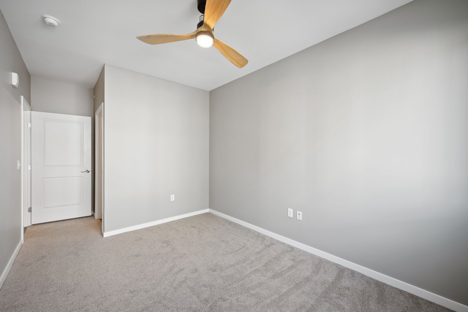 An empty room with light gray walls, carpeted flooring, a ceiling fan, and a closed white interior door.