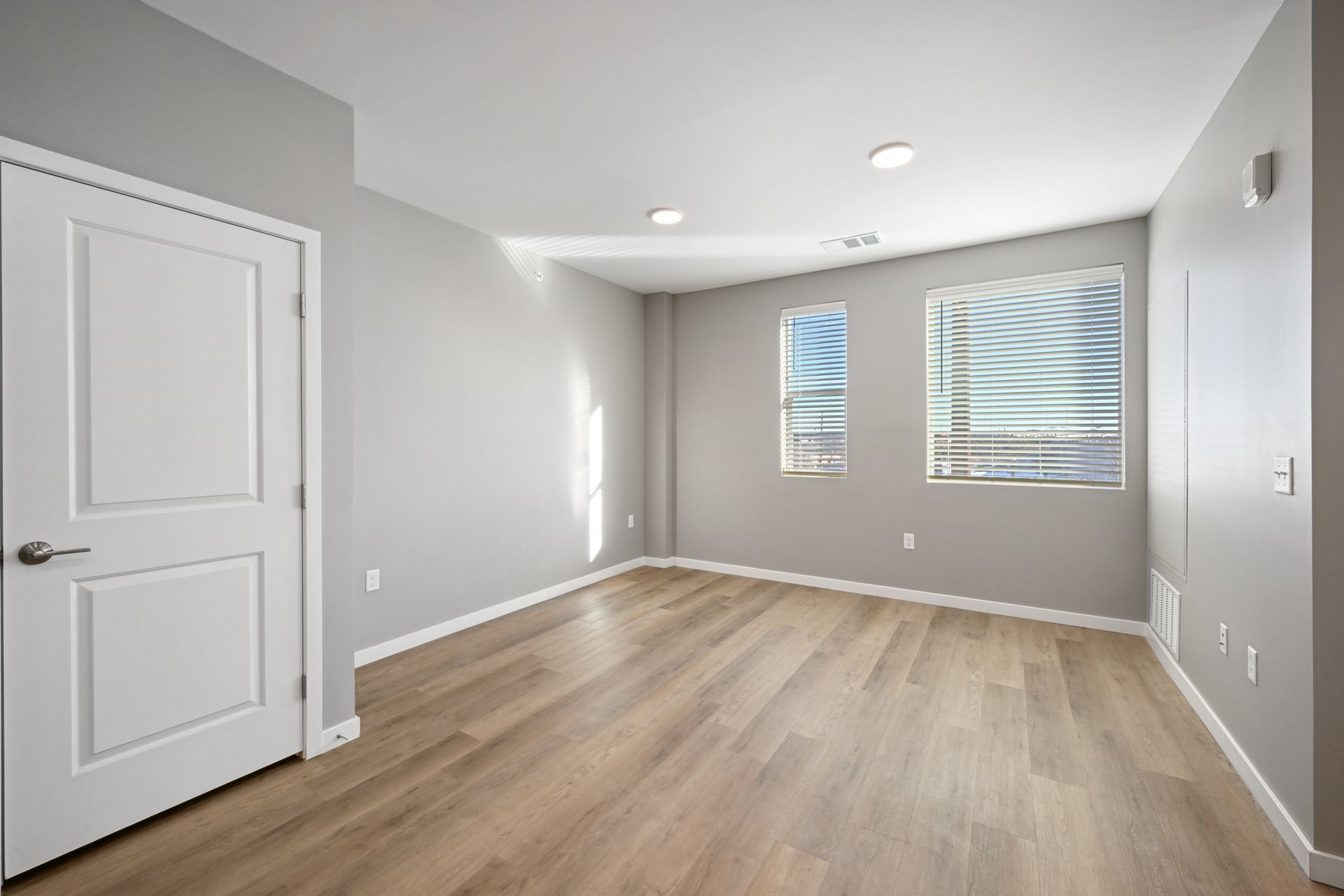A bright, empty room with light wood-style flooring, grey walls, white trim, a closed door, and two windows.