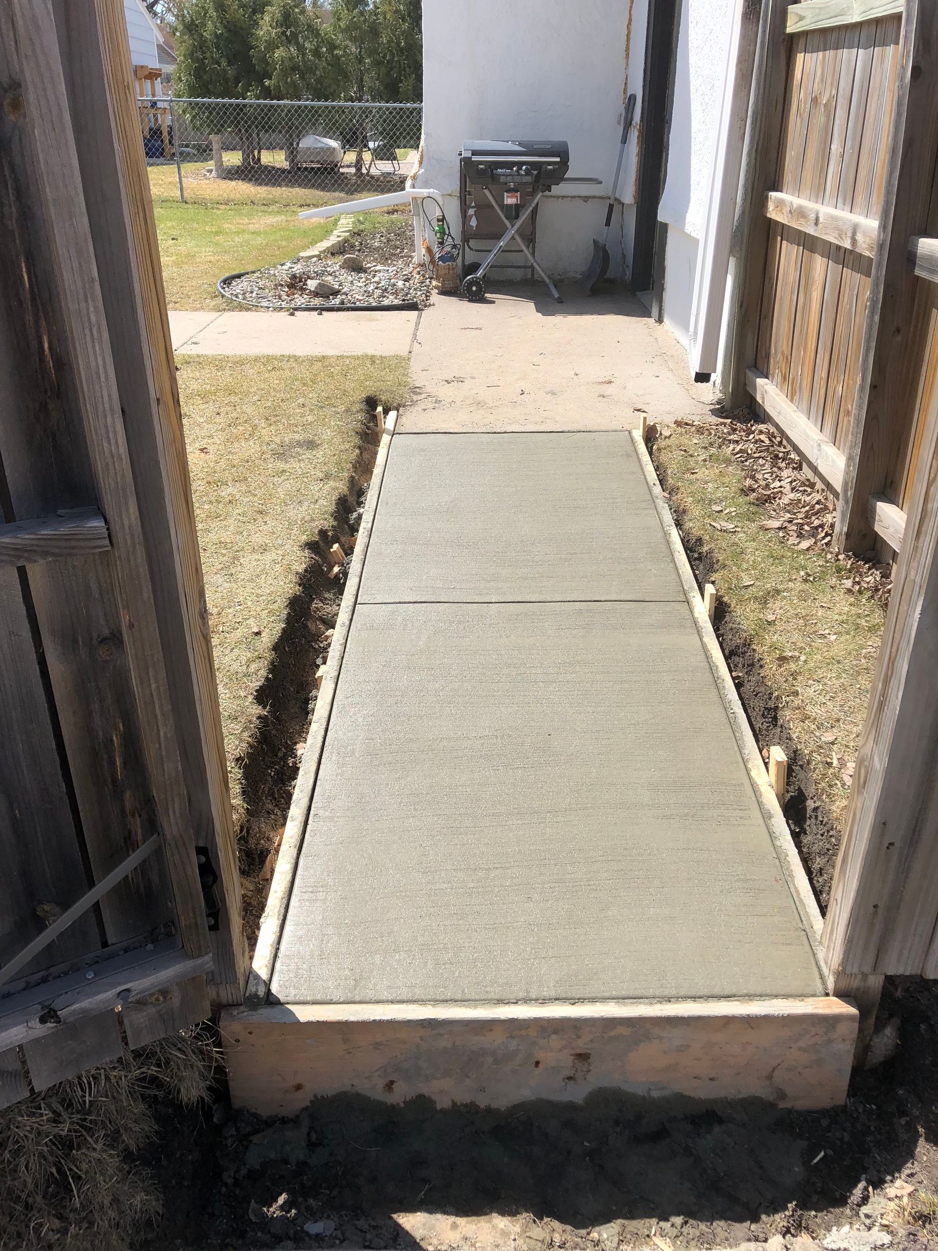 A newly poured rectangular concrete walkway sits in a narrow yard space between two wooden fences.