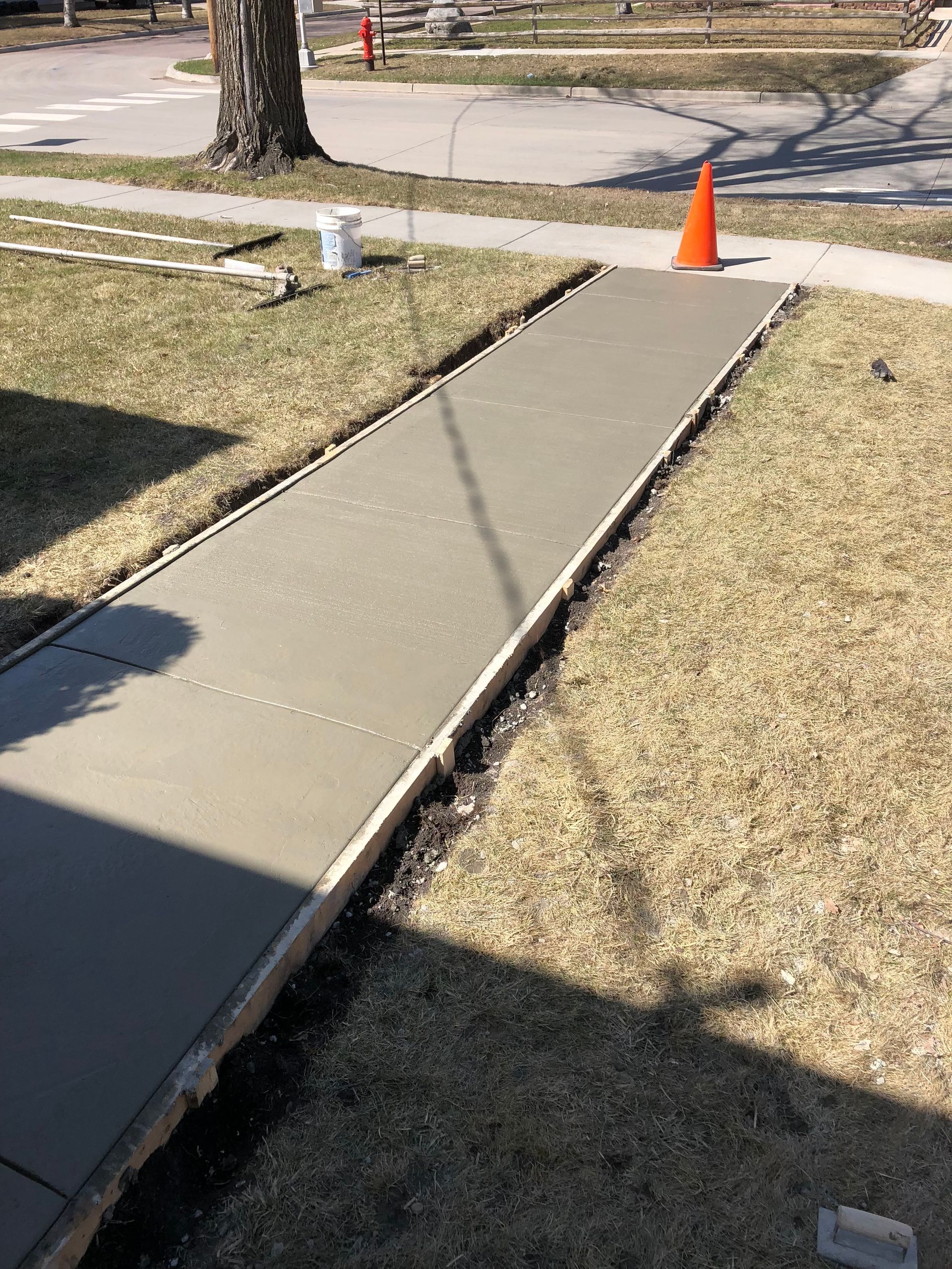 A freshly poured concrete sidewalk stretches through a grassy residential yard toward a street with an orange safety cone.