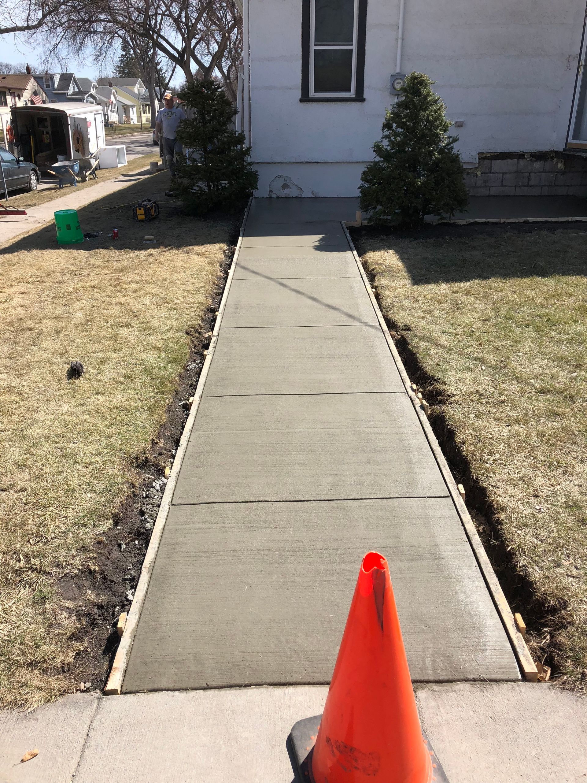 A new concrete sidewalk leads to a house, flanked by two small evergreen bushes, with an orange safety cone in front.