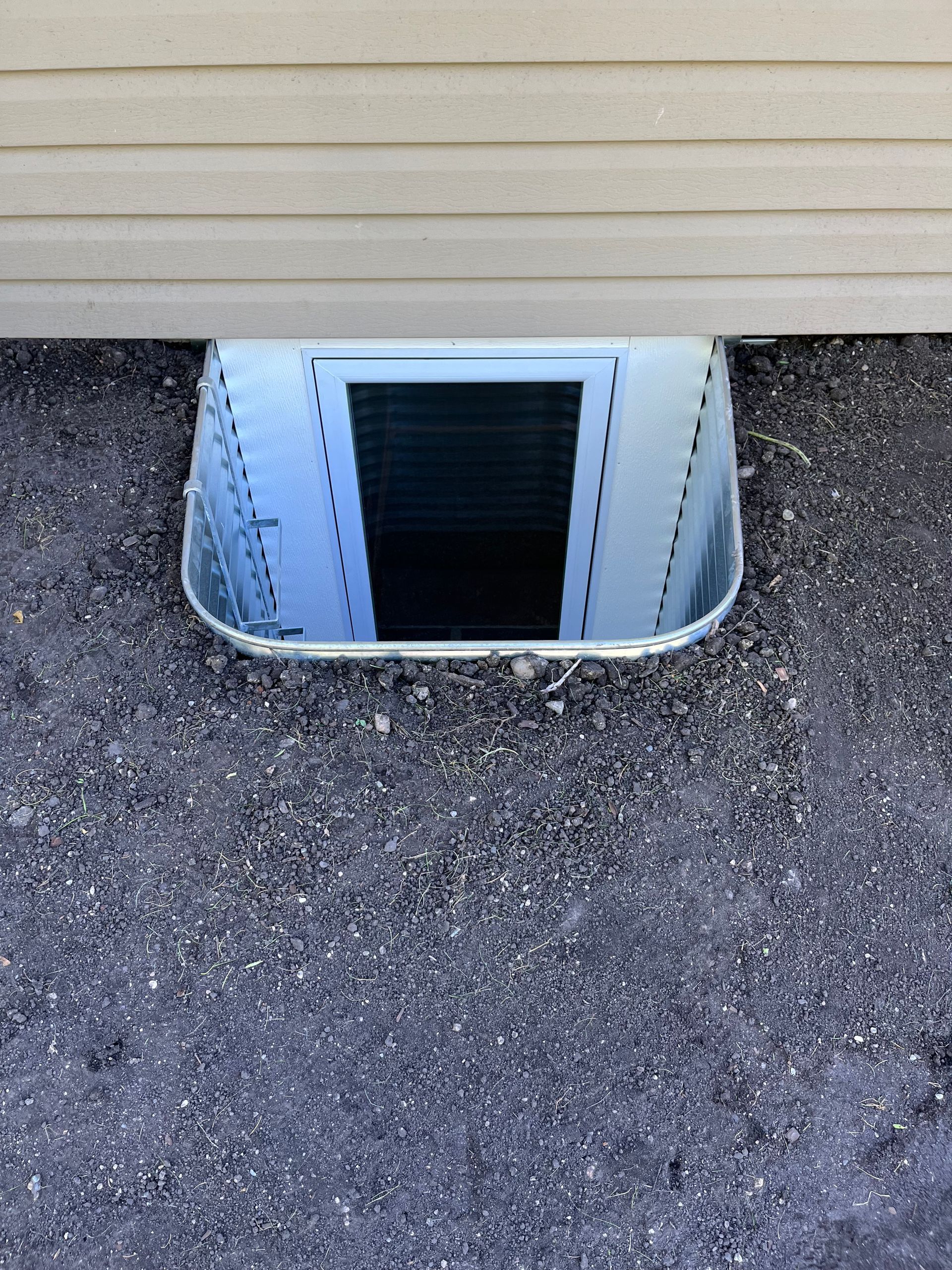 A basement window well with a metal frame and window, set into the ground against beige siding.