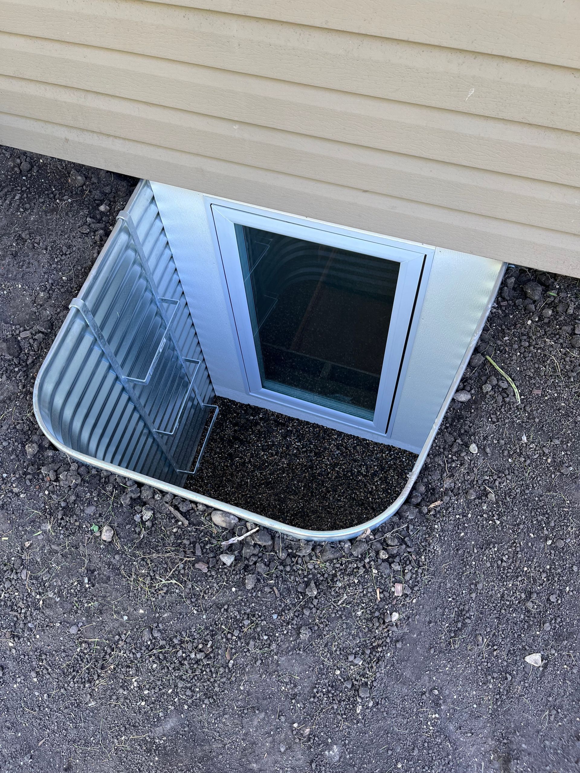 A metal basement window well installed in the ground next to a beige-sided building.