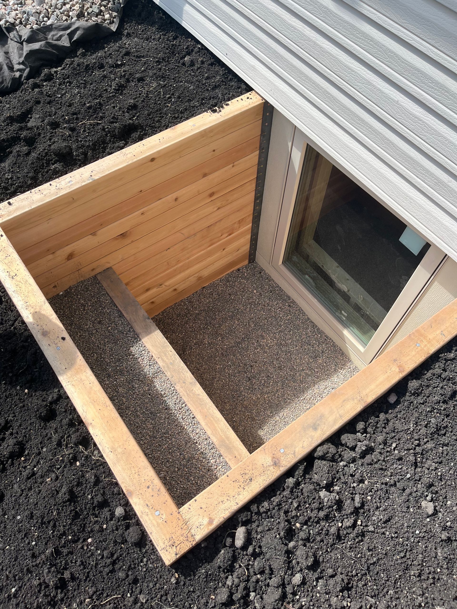 A wooden window well with an integrated staircase filled with gravel, positioned outside a basement window.