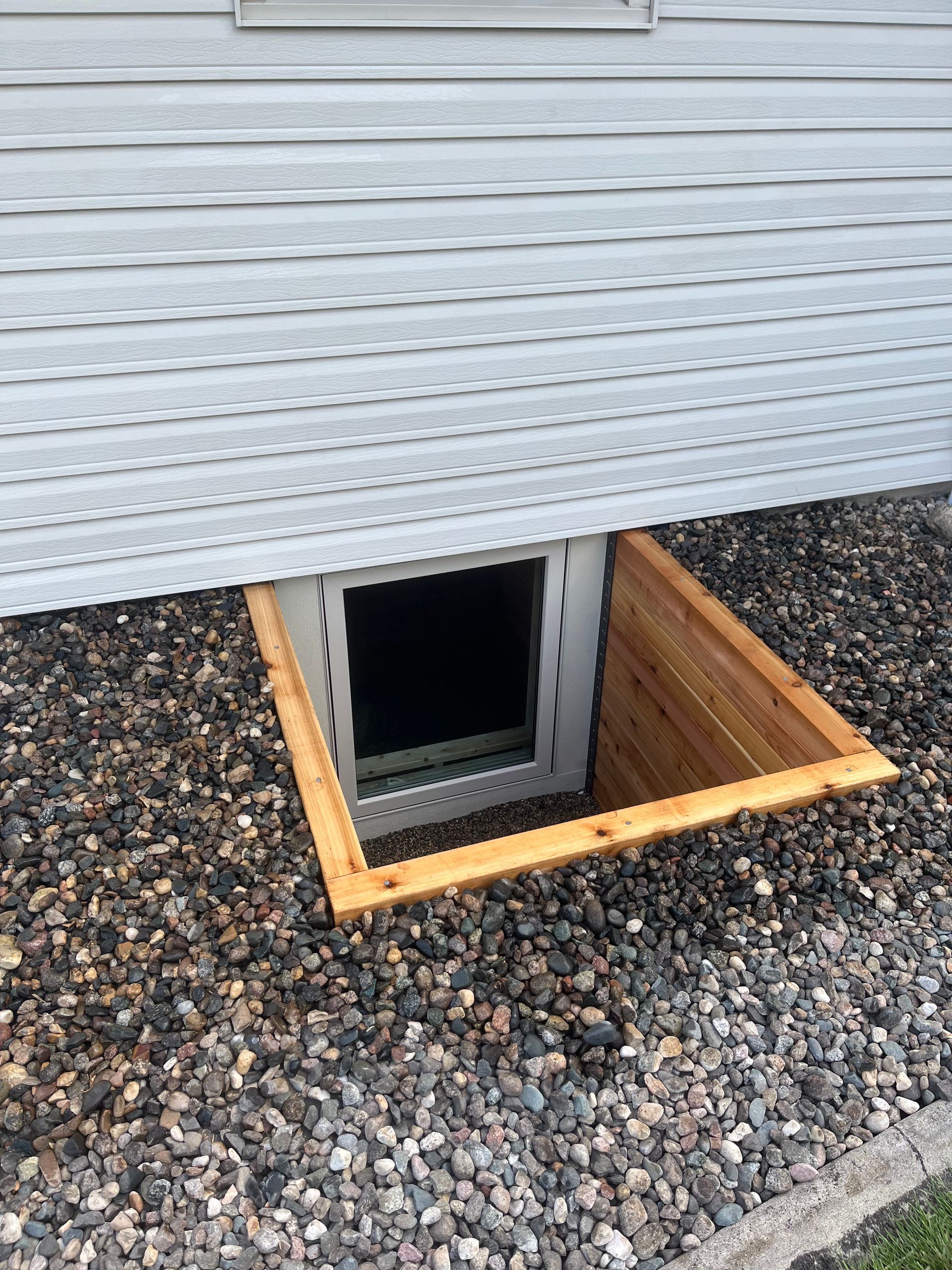 A ground-level basement window with a wooden egress well surrounded by decorative landscaping gravel.