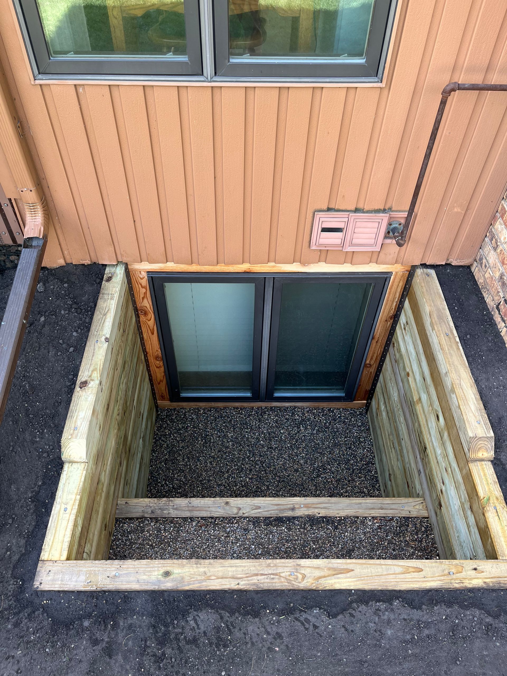A basement egress window well with wooden retaining walls and gravel fill, located below a ground-floor window.