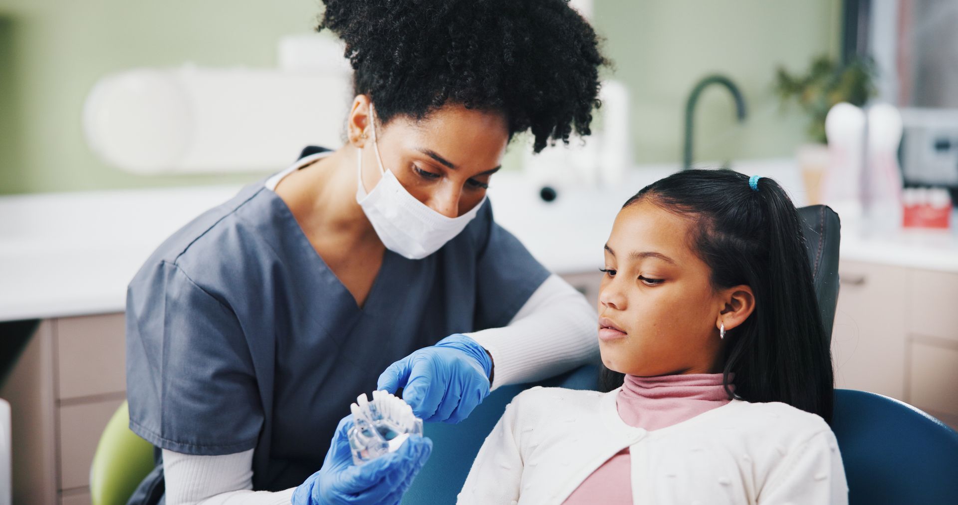 Dental hygienist in blue scrubs and mask cleans a patient’s teeth in a bright clinic
