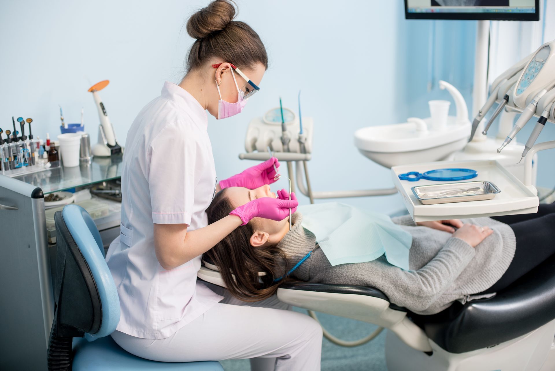 Dentist treating a reclining patient in a bright clinic with pink gloves and dental equipment nearby