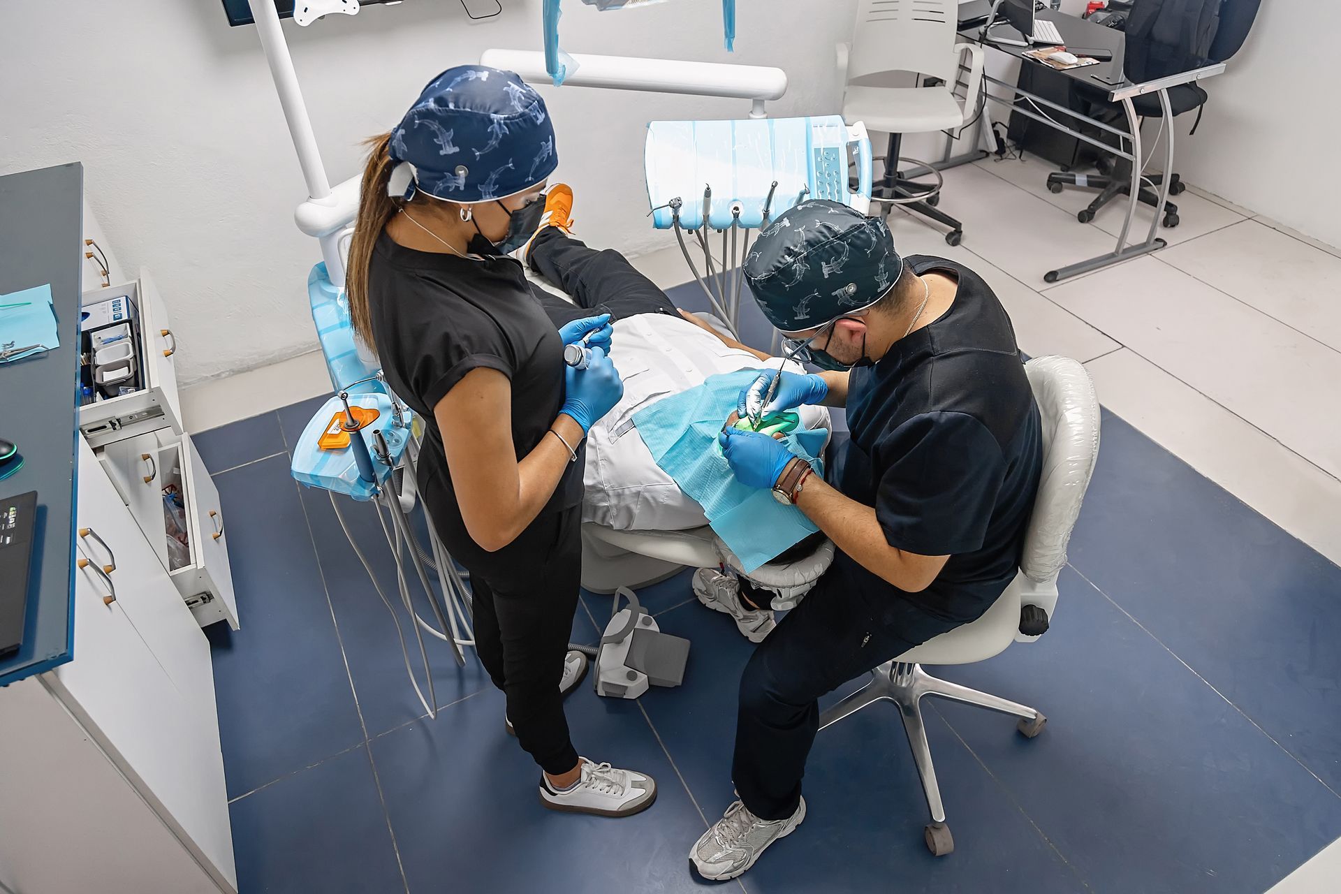 Dentists treating a patient in a dental clinic, viewed from above, with blue gowns and equipment around them