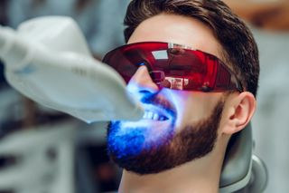 Person wearing red protective glasses receives blue dental light treatment from a dentist