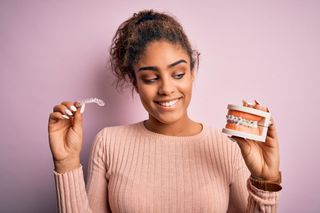 Woman holding a clear aligner and a model of teeth against a pink background