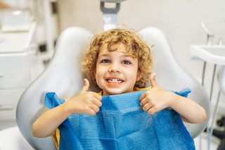 Smiling child in a dentist chair giving two thumbs up with a blue bib on