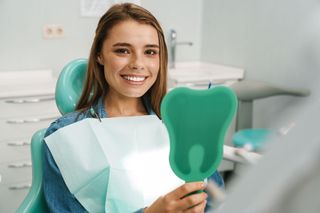 Smiling dental patient holding a green tooth-shaped mirror in a dentist’s office