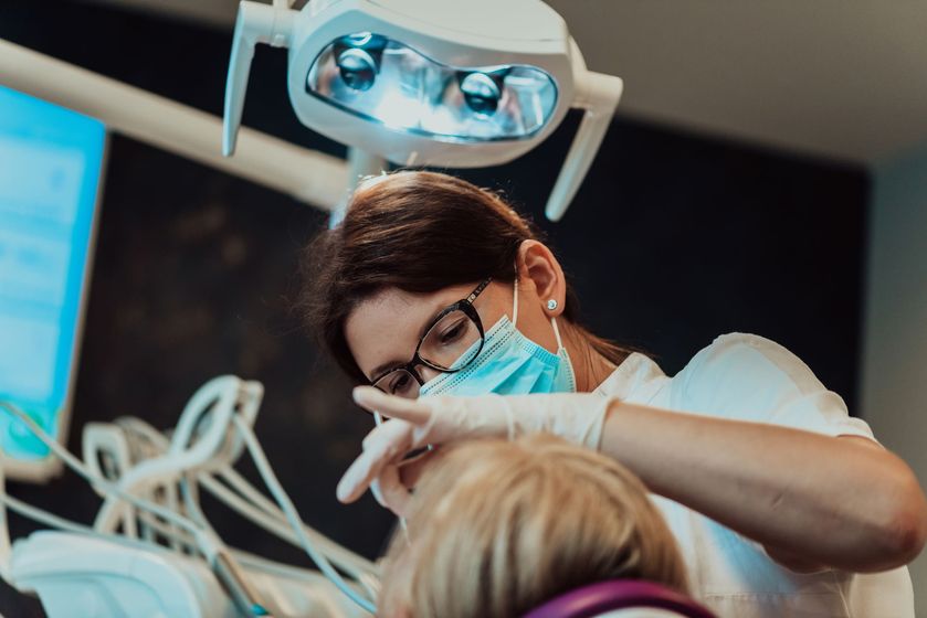 Dentist wearing a mask and glasses examines a patient under a bright overhead dental light