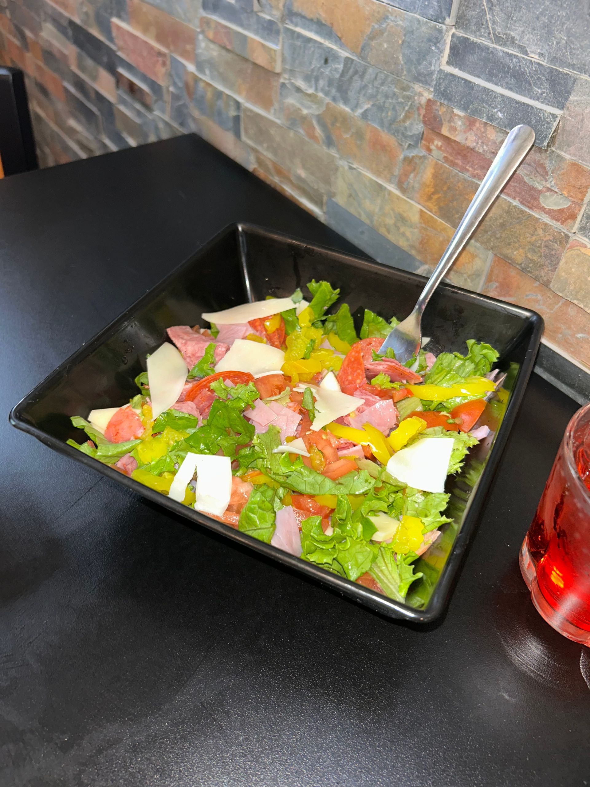 Salad in a black square bowl with lettuce, ham, tomatoes, cheese, and yellow peppers. A fork rests in the salad.