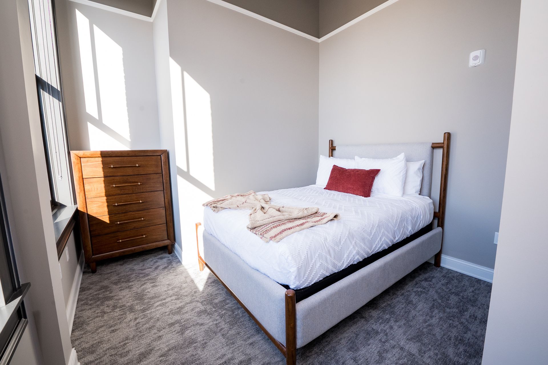 Bright bedroom with a wooden bed, white bedding, red pillow, and dresser by a sunny window.